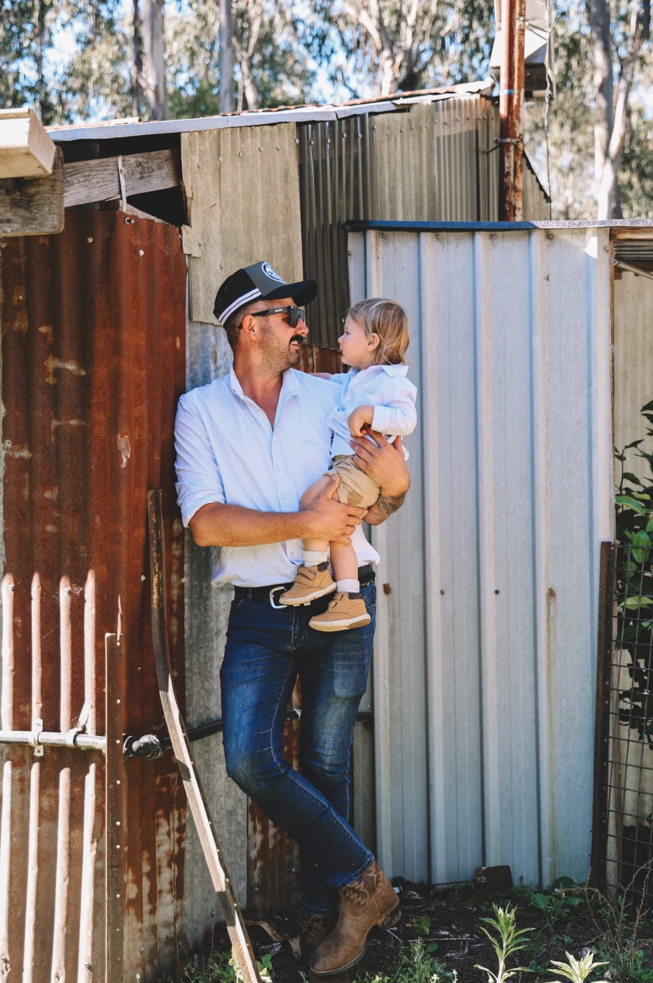 Lukas Rempel, owner of Alpine Fence, Lawn & Landscaping, holding his young son outside a shed in Myrtleford